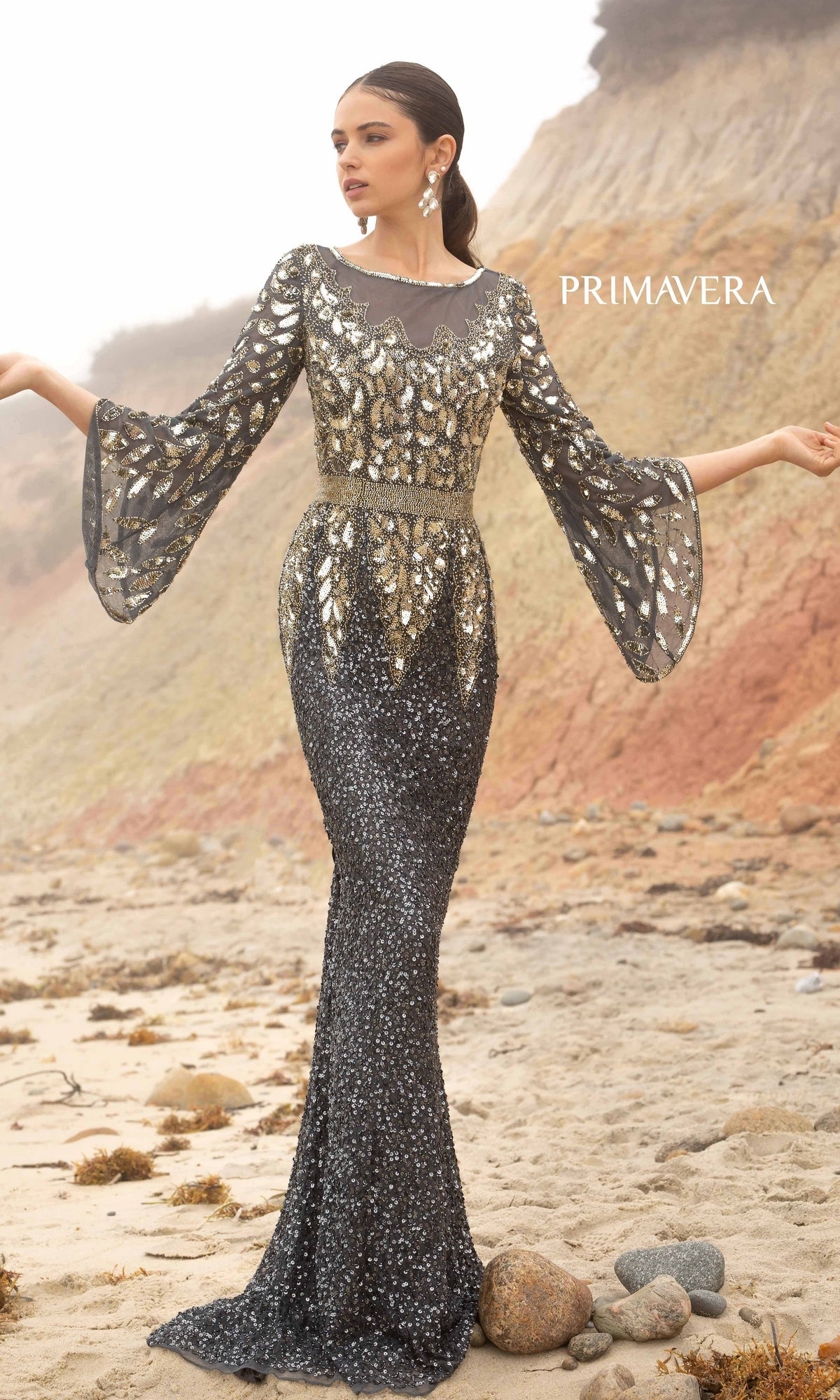 A woman in the Bell-Sleeve Long Beaded Evening Gown 1424 stands on a sandy beach, shimmering beneath cliffs and lush foliage.